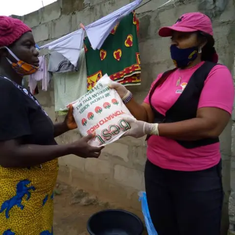 A foundation volunteer in pink hands a bag of rice to a woman wearing a burgundy crochet-style hat at the food distribution event.