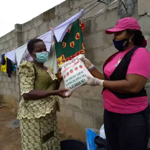 A foundation volunteer in pink hands a bag of rice to a woman in a green dress at an outdoor food distribution.