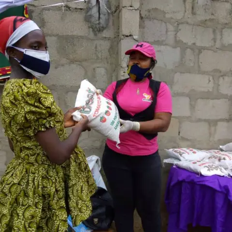A foundation volunteer in pink hands a bag of rice to a young woman in a green printed dress, with the AFSF banner visible behind them.