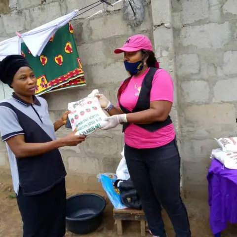 A foundation volunteer in pink hands a bag of rice to a woman in a grey and white checked top at an outdoor distribution event.