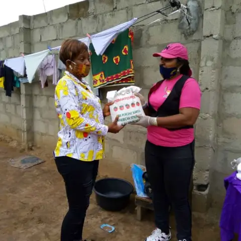 A foundation volunteer in pink hands a bag of Best Tomato Rice to a woman in a yellow floral blouse during a door-to-door food distribution.