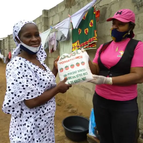 A foundation volunteer in pink hands a bag of rice to a woman in a white polka-dot dress at an outdoor food distribution.