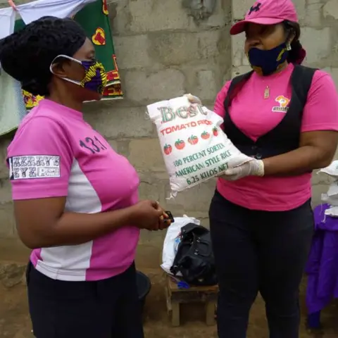 A foundation volunteer in pink hands a bag of rice to a woman in a pink and black sports shirt at an outdoor food distribution event.