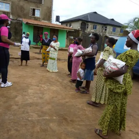A line of community women wearing face masks, each holding a bag of rice received during an outdoor food distribution event.