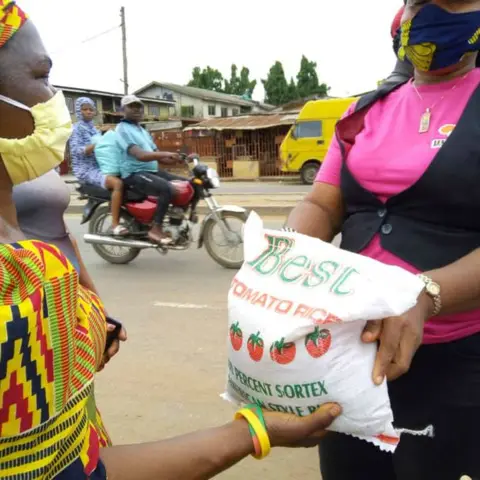 A foundation volunteer in pink hands a bag of rice to a woman in a kente-print dress at a busy street corner, with a motorcycle passing in the background.