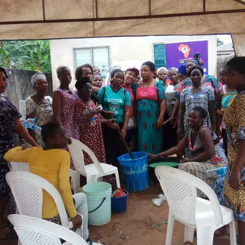 Women standing around large buckets of green liquid soap produced during a soap-making empowerment workshop under a canopy tent.