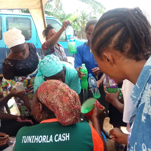 Women hold up bottles of liquid soap they made during an outdoor vocational skills training session.
