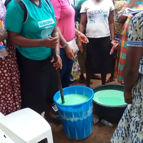 A woman in a teal Igidaduro t-shirt stirs large buckets of green liquid soap as other women observe during an empowerment skills workshop.