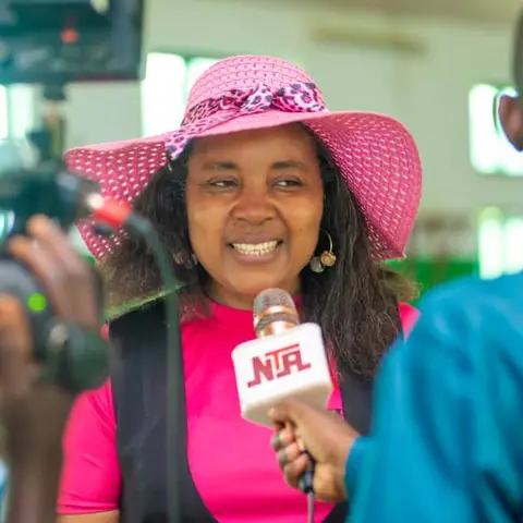 A woman in a pink wide-brimmed hat smiling during an NTA television interview at an outdoor community event.