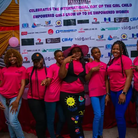 Seven foundation team members in pink LFAGCW shirts posing in front of the International Day of the Girl Child banner with the theme 'Empowered Girls Grow Up To Be Empowered Women.'
