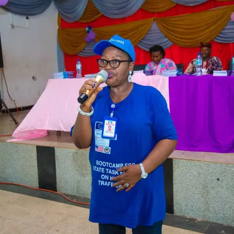 A woman in a blue NAPTIP Bootcamp t-shirt and cap speaks into a microphone on a decorated stage at the International Day of the Girl Child event.