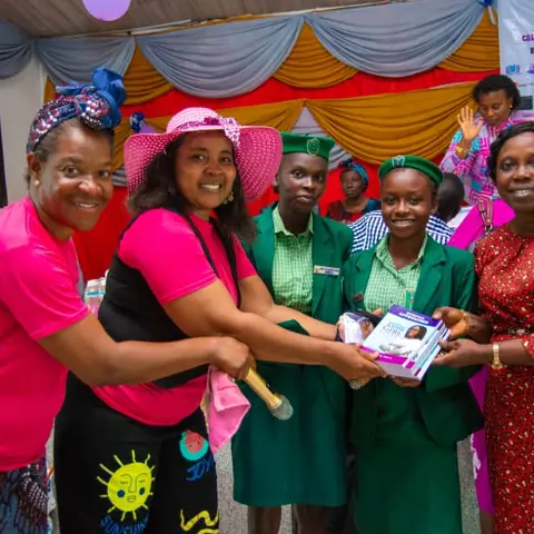 Foundation volunteers in pink present sanitary products and books to two schoolgirls in green uniforms at the International Day of the Girl Child celebration.