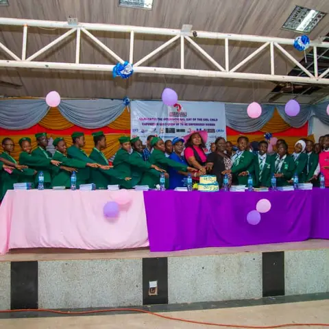 Students and foundation organisers on a colourfully decorated stage cutting a cake together at the International Day of the Girl Child celebration, with school pupils lined along a table.