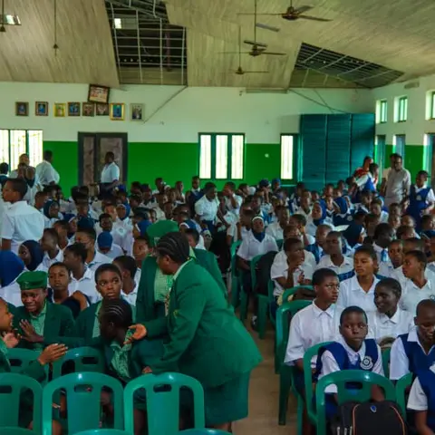 A crowded school hall full of pupils in green and blue uniforms, with a teacher leaning in to speak to a student in the front row during the Girl Child Day event.