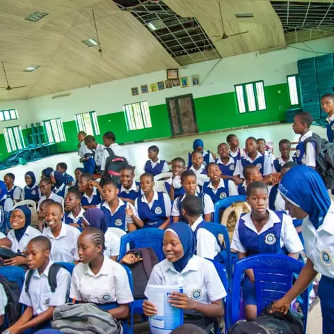 Schoolgirls in blue and white uniforms chatting and smiling while seated in blue plastic chairs in a school hall.