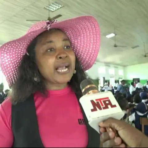 A close-up of a woman in a pink hat being interviewed by NTA television in a school hall during the Girl Child Day event.