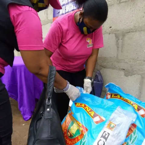 Two foundation volunteers in pink shirts and gloves sort bags of rice beside the African Girl Child and Women banner during a food distribution event.