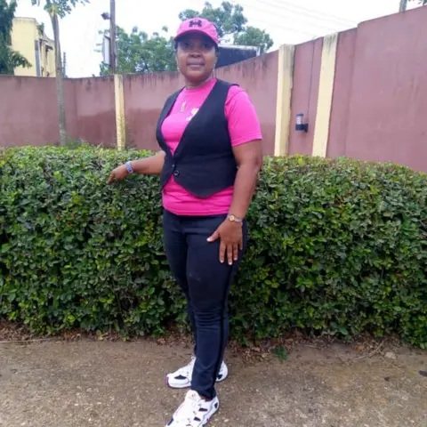 A foundation volunteer in a pink t-shirt and cap poses outside by a garden hedge before the community outreach programme.