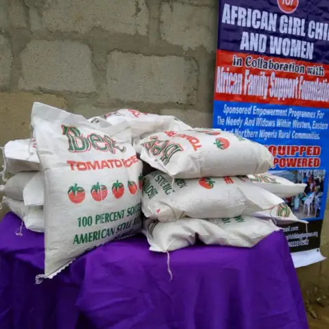 Bags of Best Tomato Rice stacked on a purple table in front of the African Family Support Foundation and AGCW banner, prepared for community distribution.