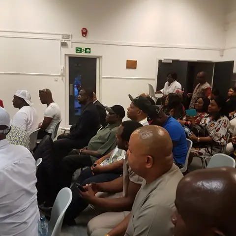 A woman in a peach top speaking to a seated audience in a formal room.