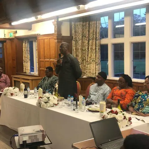 A man in a traditional patterned shirt speaking to an audience in a formal conference room with a projector screen.