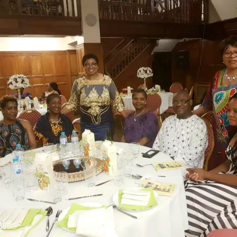 A woman in a teal shirt stirring a large tub of green liquid under a tent, with other women seated nearby.