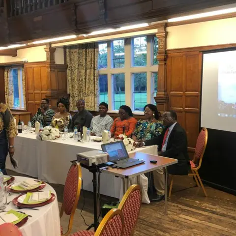 A group photo of people in formal and traditional wear in a wood-paneled hall.