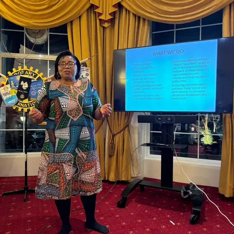 A woman in a vibrant, colorful patterned dress giving a presentation in front of a TV screen titled 'WHAT WE DO,' listing services like family mediation and parenting strategies.