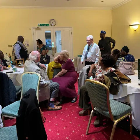A man in a white shirt and tie stands among seated guests at a round table during a break or transition in the presentation.