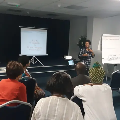 A presenter speaking next to a projector screen titled 'DA/DV Complexities in African Immigrant Communities' during a workshop.