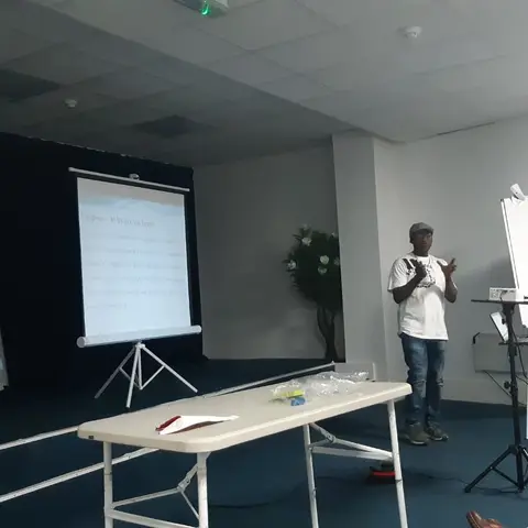 A man gesturing while giving a presentation next to a whiteboard and projector screen in a meeting room.