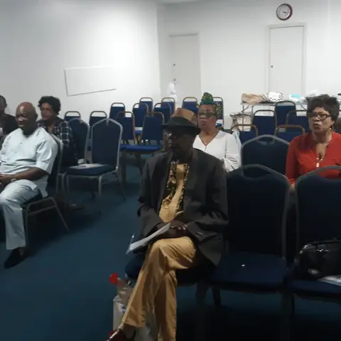 An audience seated in blue chairs listening attentively to a presentation in a community hall.