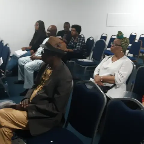 A side view of attendees seated in rows, focused on the speaker at the front of the room.