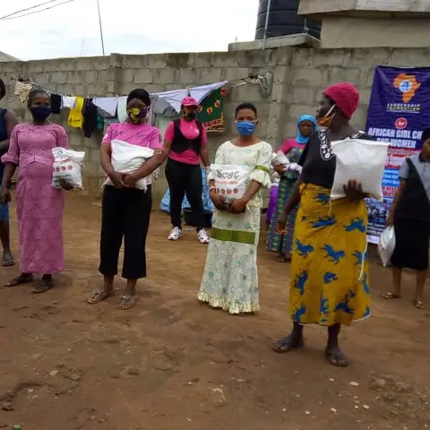 Community women wearing face masks stand in a socially-distanced line, each holding a bag of rice received from the foundation, with the LFAGCW banner visible.
