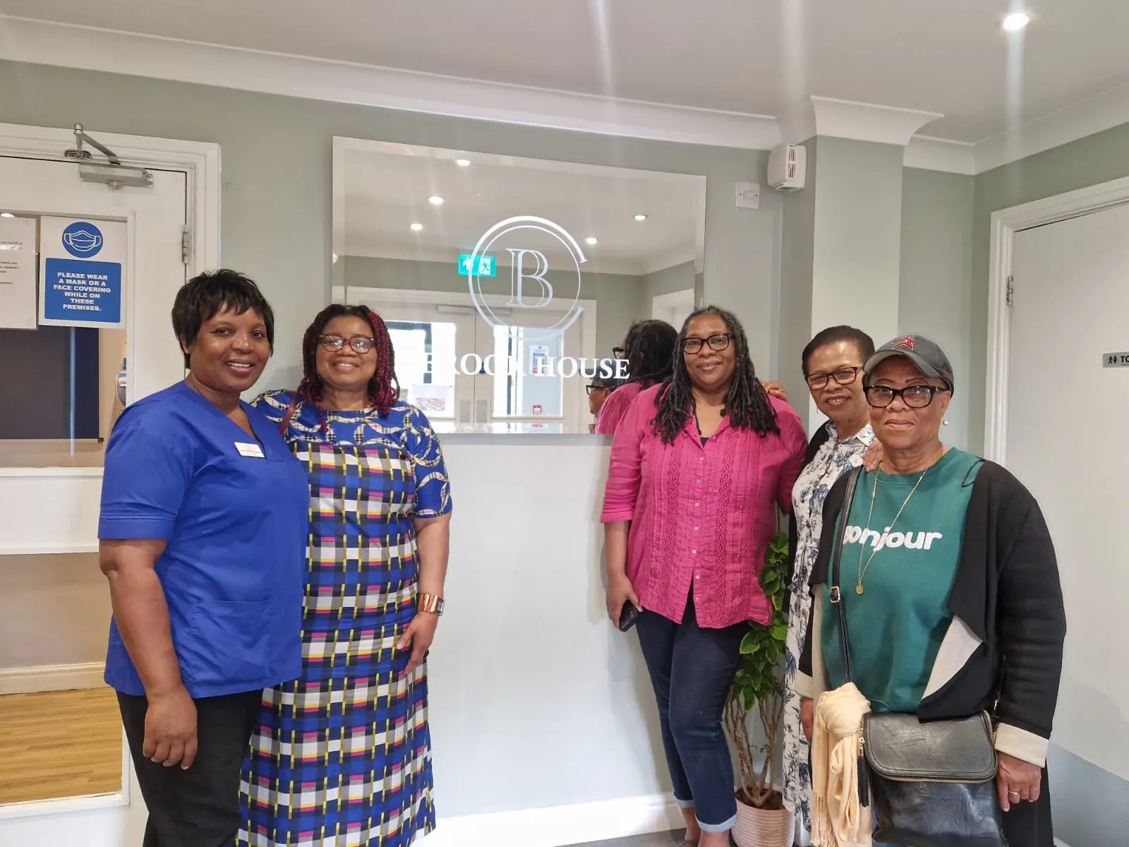 Five women standing together in the lobby of Brook House, smiling for a group photo. One woman is wearing blue medical scrubs.