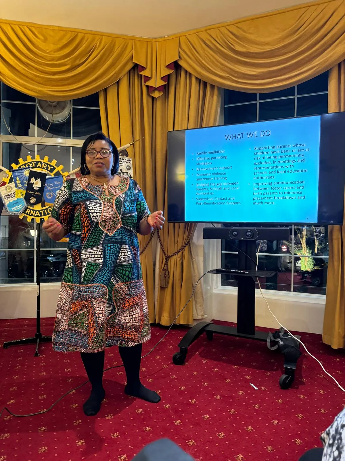 A woman in a vibrant, colorful patterned dress giving a presentation in front of a TV screen titled 'WHAT WE DO,' listing services like family mediation and parenting strategies.